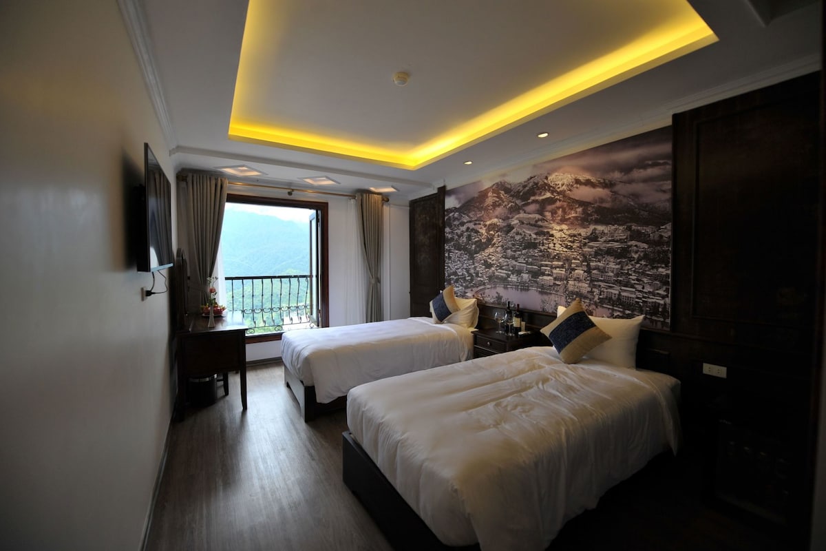 A hotel room with two beds, a desk, a view of the mountains, and a cityscape mural.