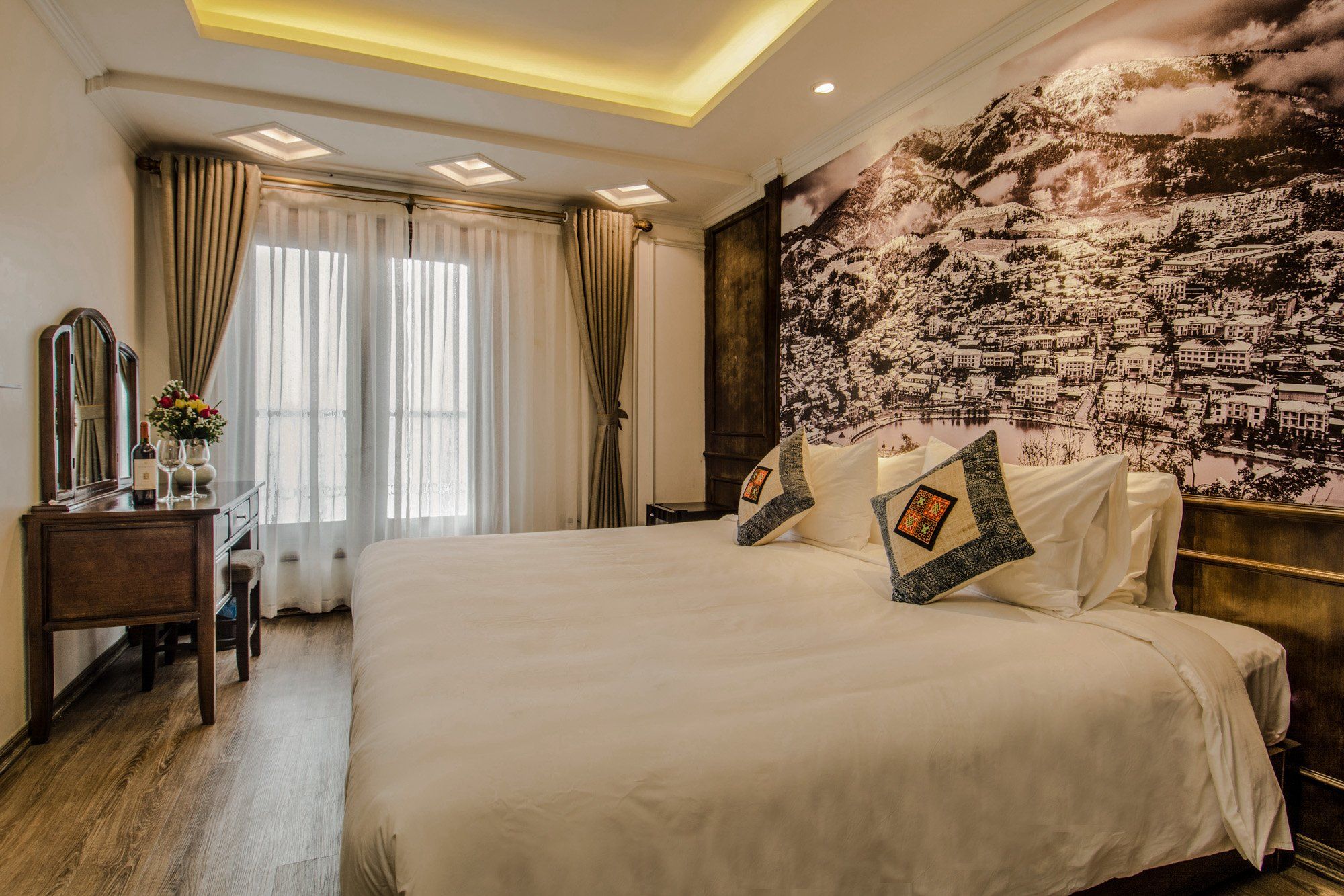 A hotel room with a large bed, a desk with a mirror and a vase of flowers, and a cityscape mural on the wall.