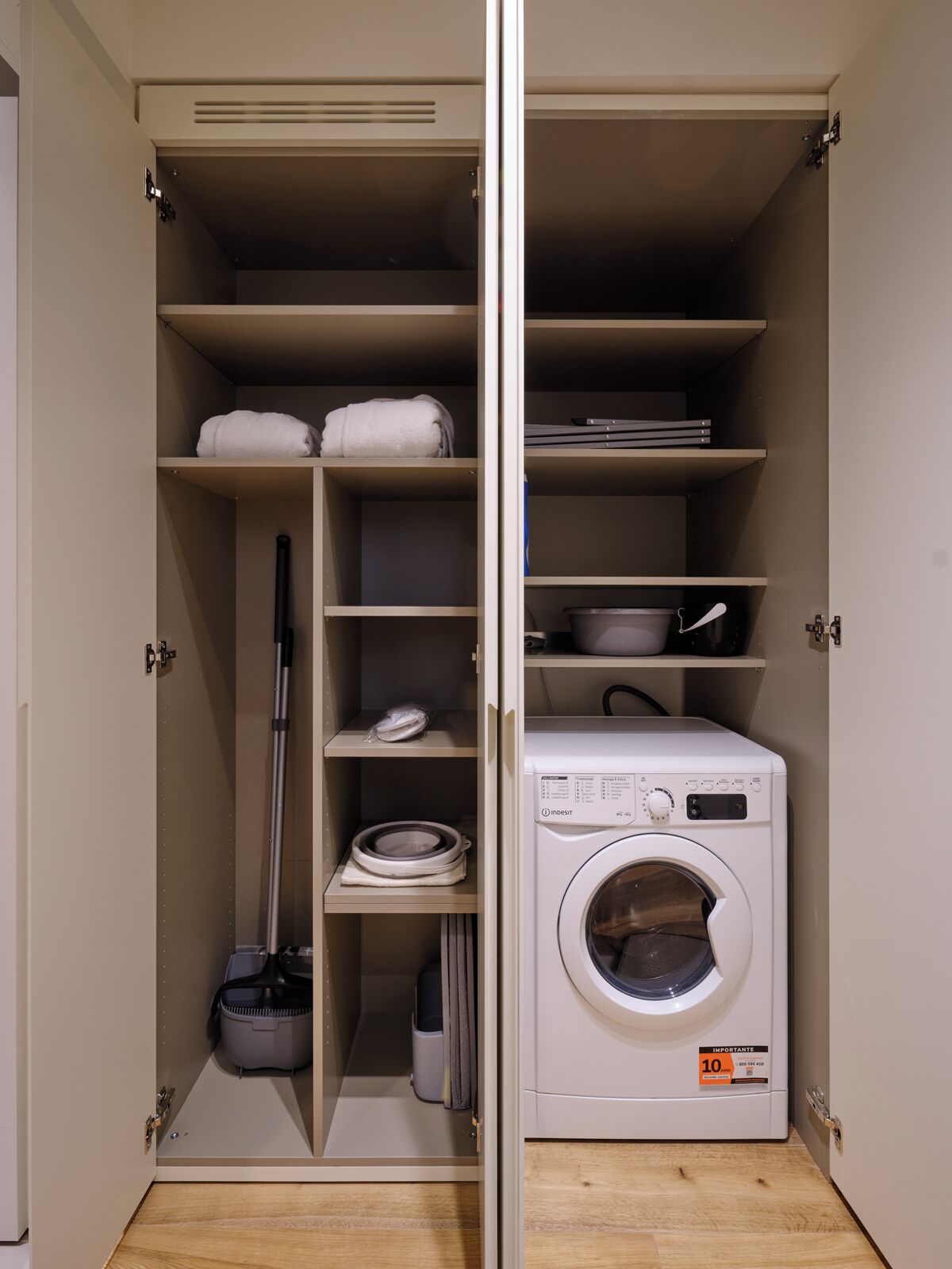 A laundry room with a front-loading washing machine, shelves with folded towels, and a mop.