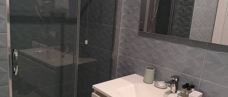 Un baño moderno con cabina de ducha de vidrio, un lavamanos blanco con un expositor de artículos de aseo y un espejo grande sobre el lavamanos.