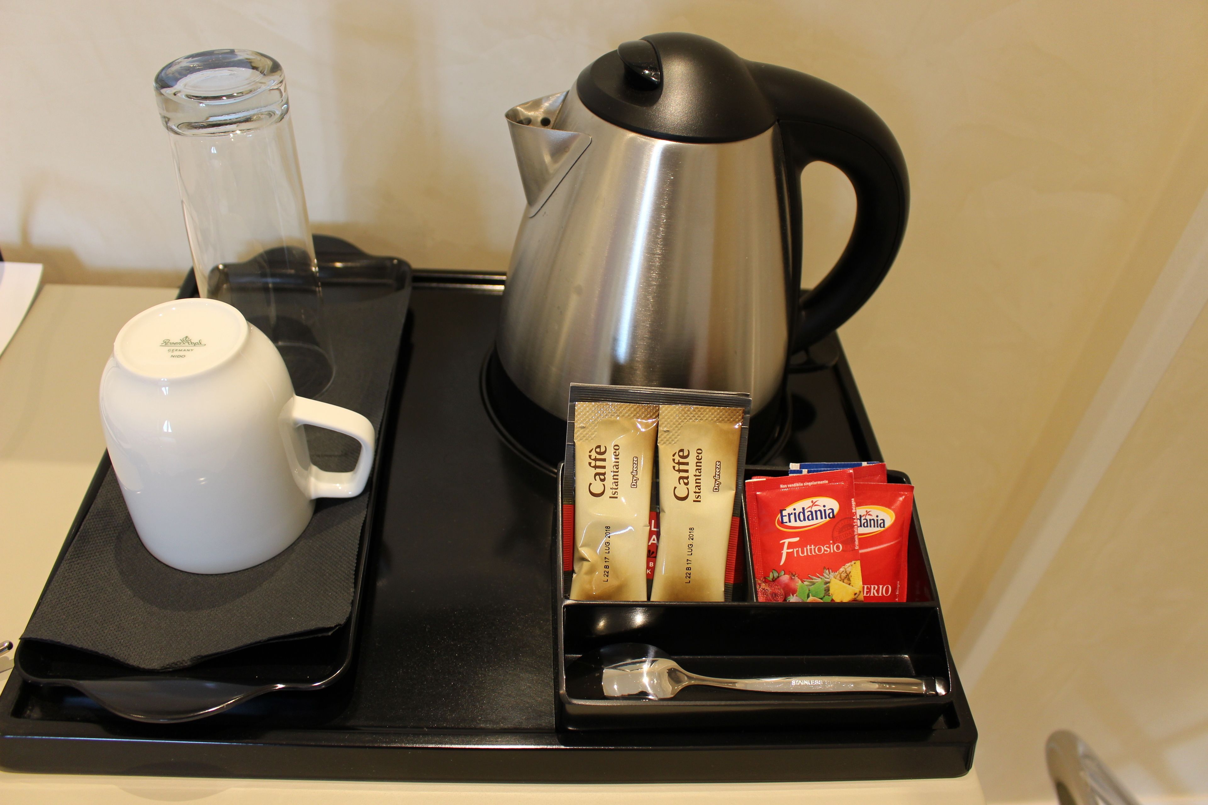 Un plateau avec une tasse blanche, un verre, une bouilloire en acier inoxydable, des dosettes de café espresso Caffe Italia, un sachet de Fruttosio et une cuillère.