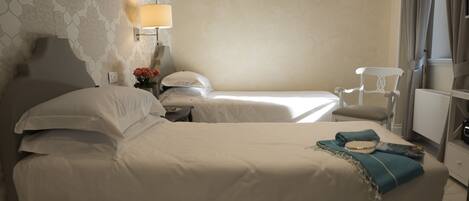 Quarto de hotel com duas camas, mesa de cabeceira com luminária, uma cadeira e janela com cortinas.