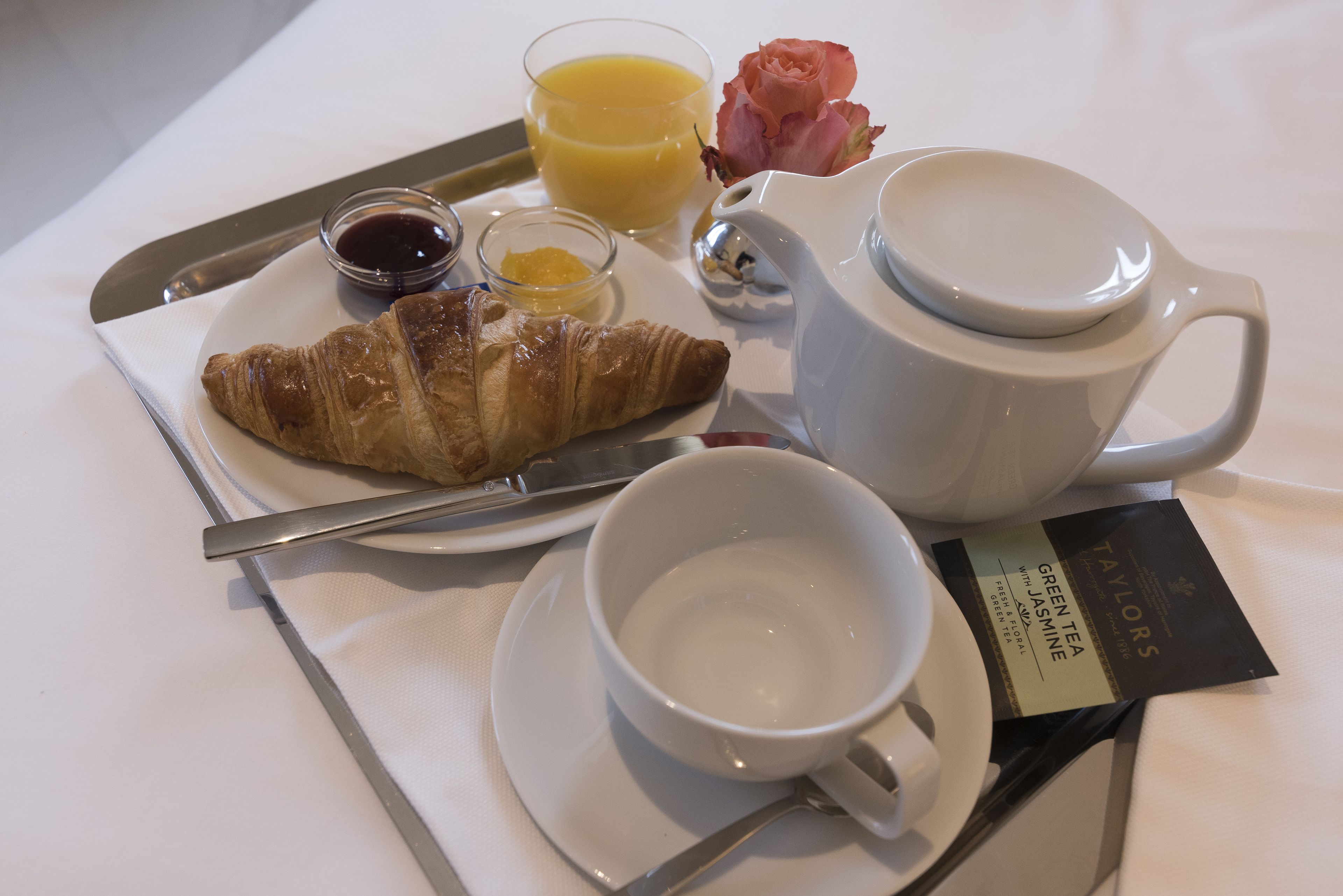 A tray with a croissant, a glass of orange juice, a small bowl of jam, a cup of tea, and a rose.