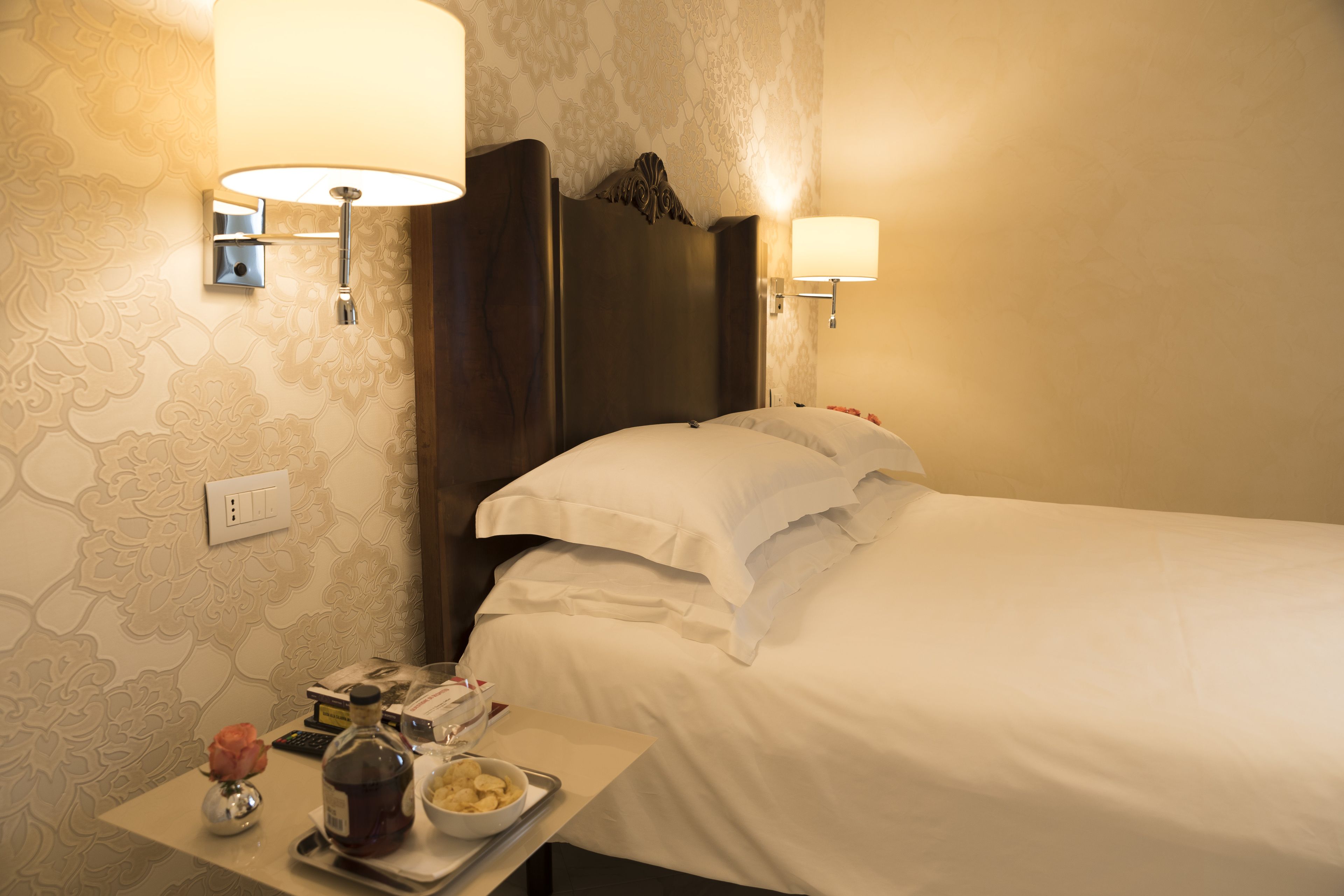 Quarto de hotel com uma cama, duas luminárias na parede, uma mesinha de cabeceira com um vaso, uma garrafa e uma tigela.