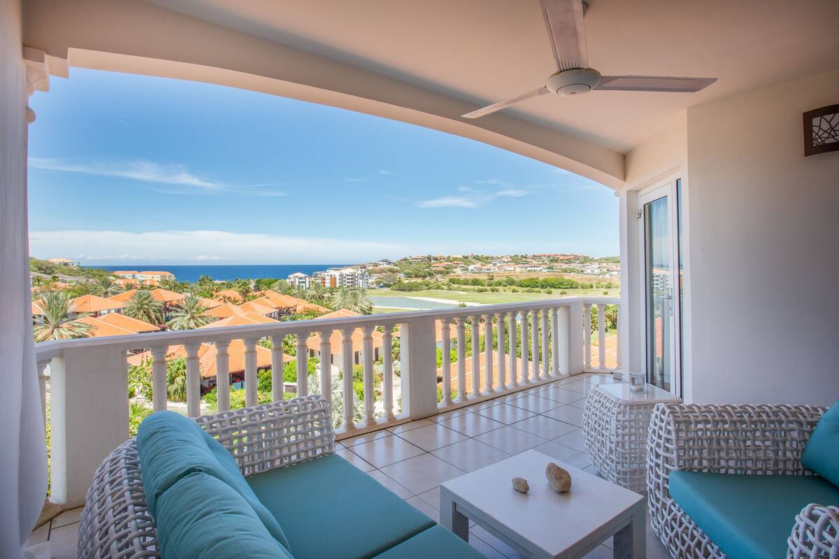 A balcony with white wicker furniture, a ceiling fan, and a view of the sea and buildings.