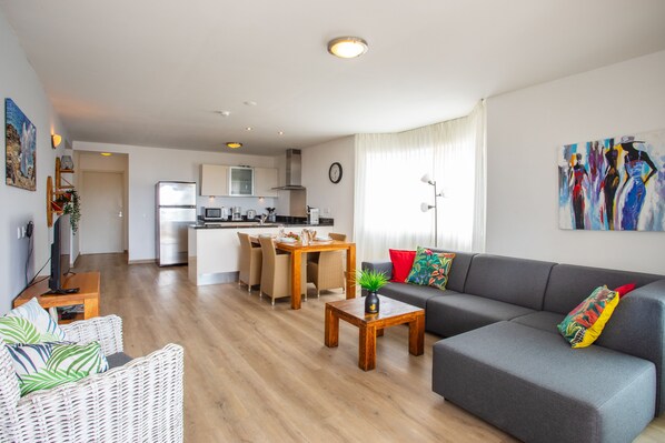 A modern living room with a grey sofa, a wooden coffee table, and a dining area with a kitchen in the background.