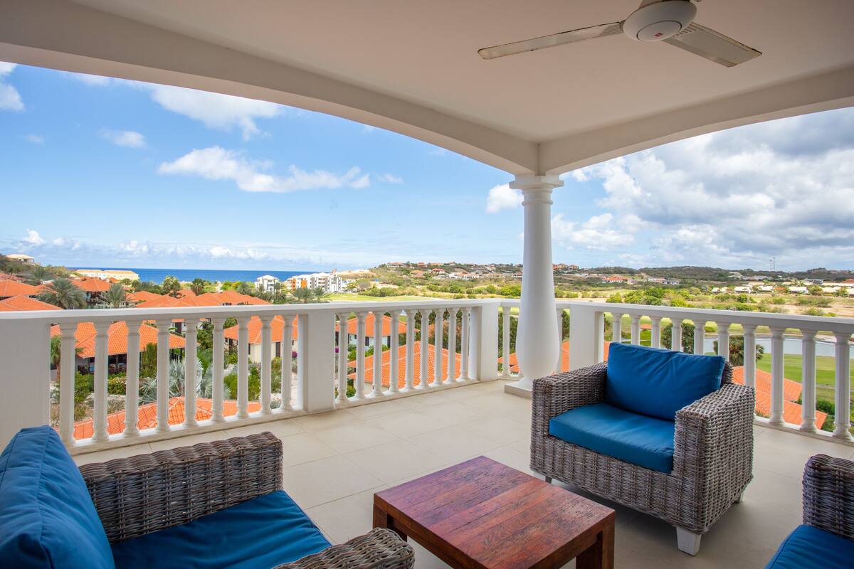 A balcony with wicker furniture, a ceiling fan, and a view of a coastal town.