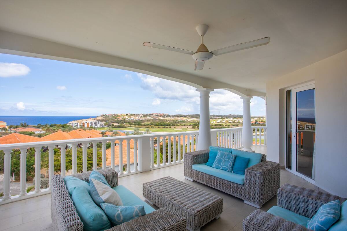 A balcony with wicker furniture, a ceiling fan, and a view of the sea and buildings.