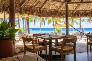 An outdoor seating area with wooden tables and chairs, overlooking a beach with palm trees and the ocean. - Blue Bay Bungalows (Sint Michiel)
