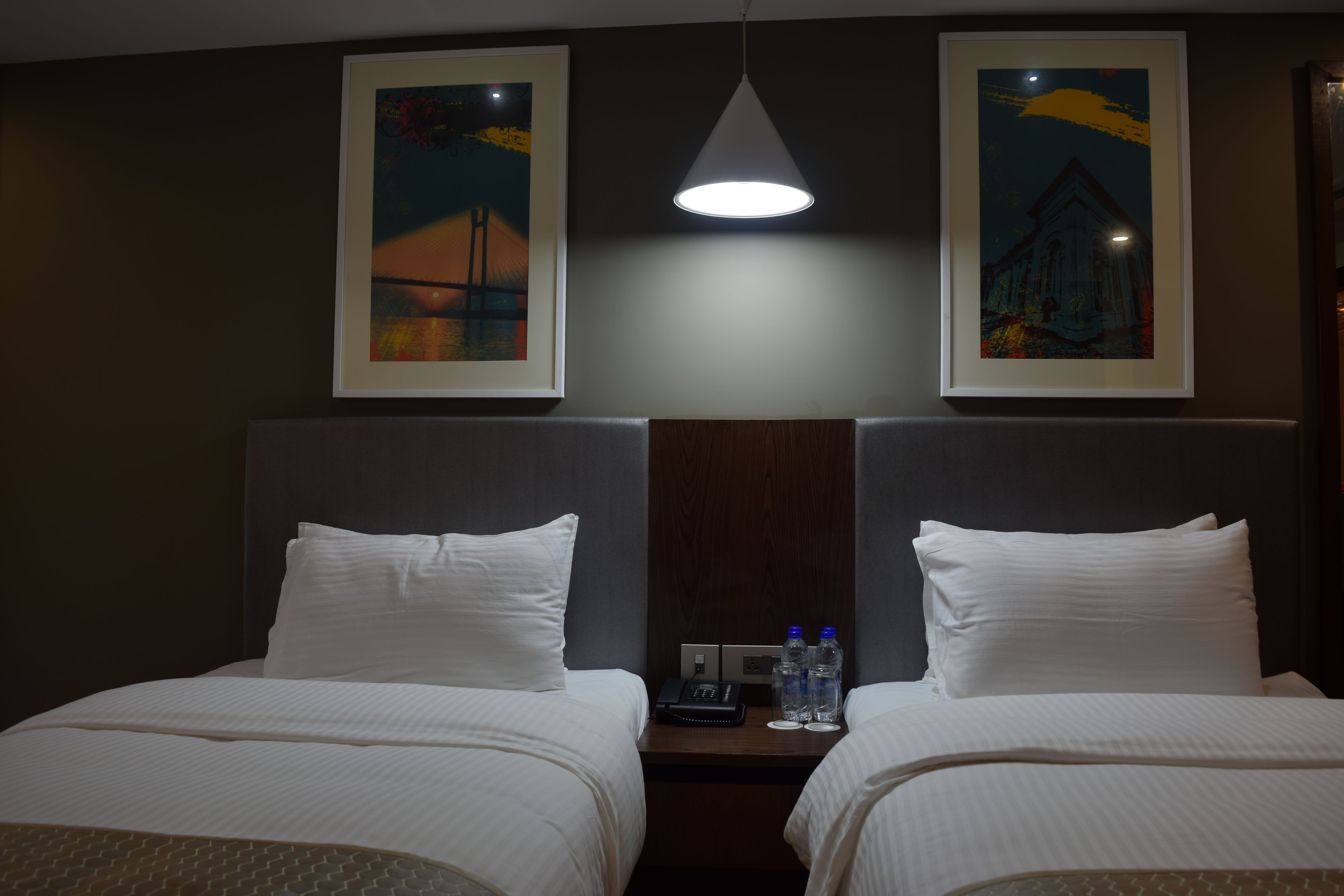 Une chambre d’hôtel avec deux lits, une table de chevet avec un téléphone et des bouteilles d’eau, et des œuvres d’art encadrées accrochées aux murs.