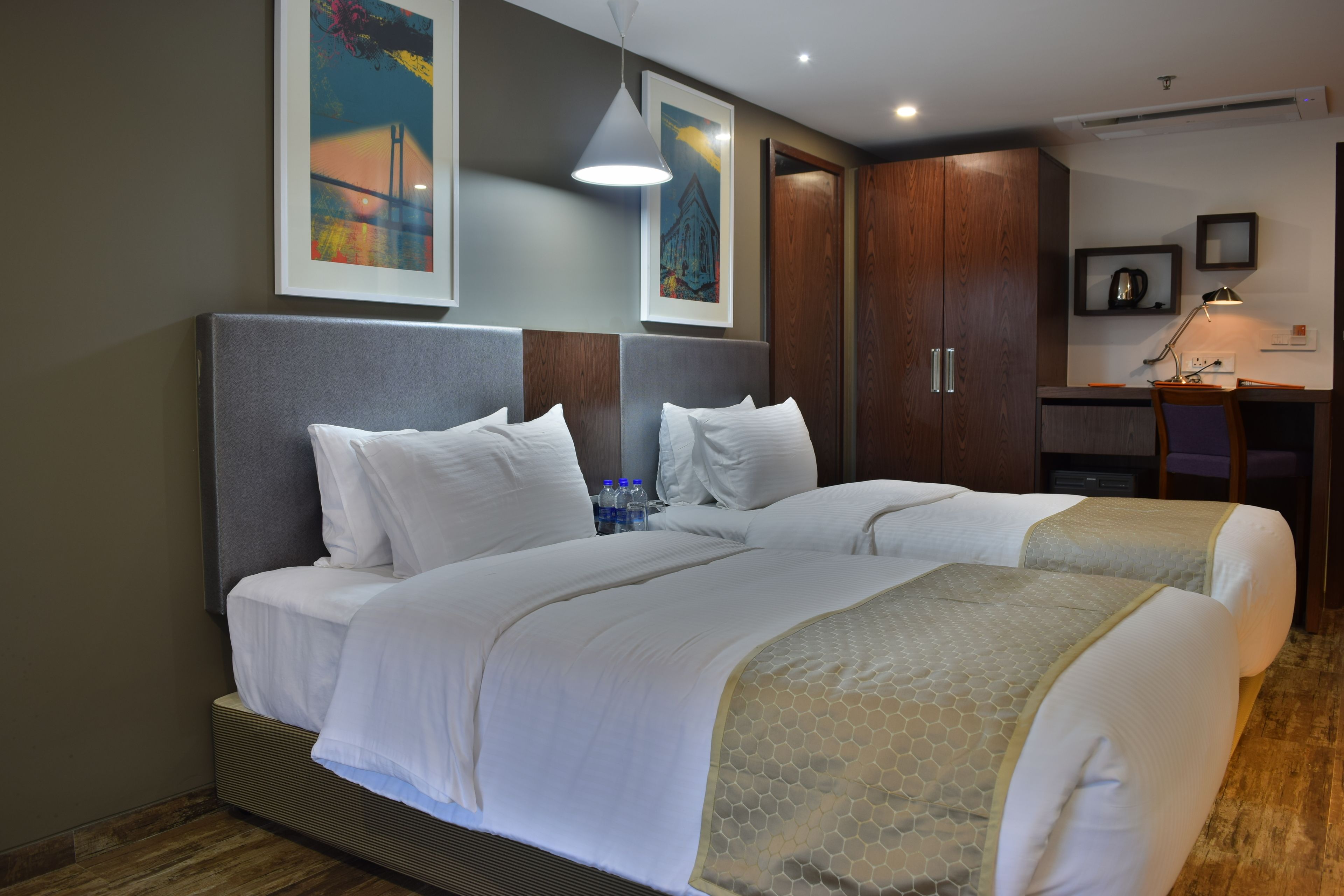 A hotel room with two beds, a desk, and a chair. There are framed pictures on the wall, a ceiling light, and a window with a view of a building.