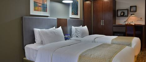 A hotel room with two beds, a desk, and a chair. There are framed pictures on the wall, a ceiling light, and a window with a view of a building.