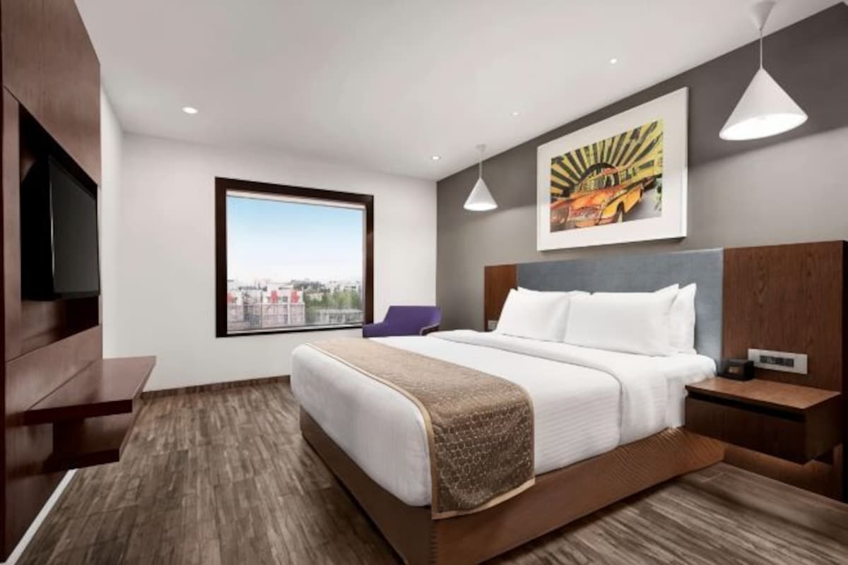 A modern hotel room with a large bed, a television, a window with a city view, and a painting on the wall.