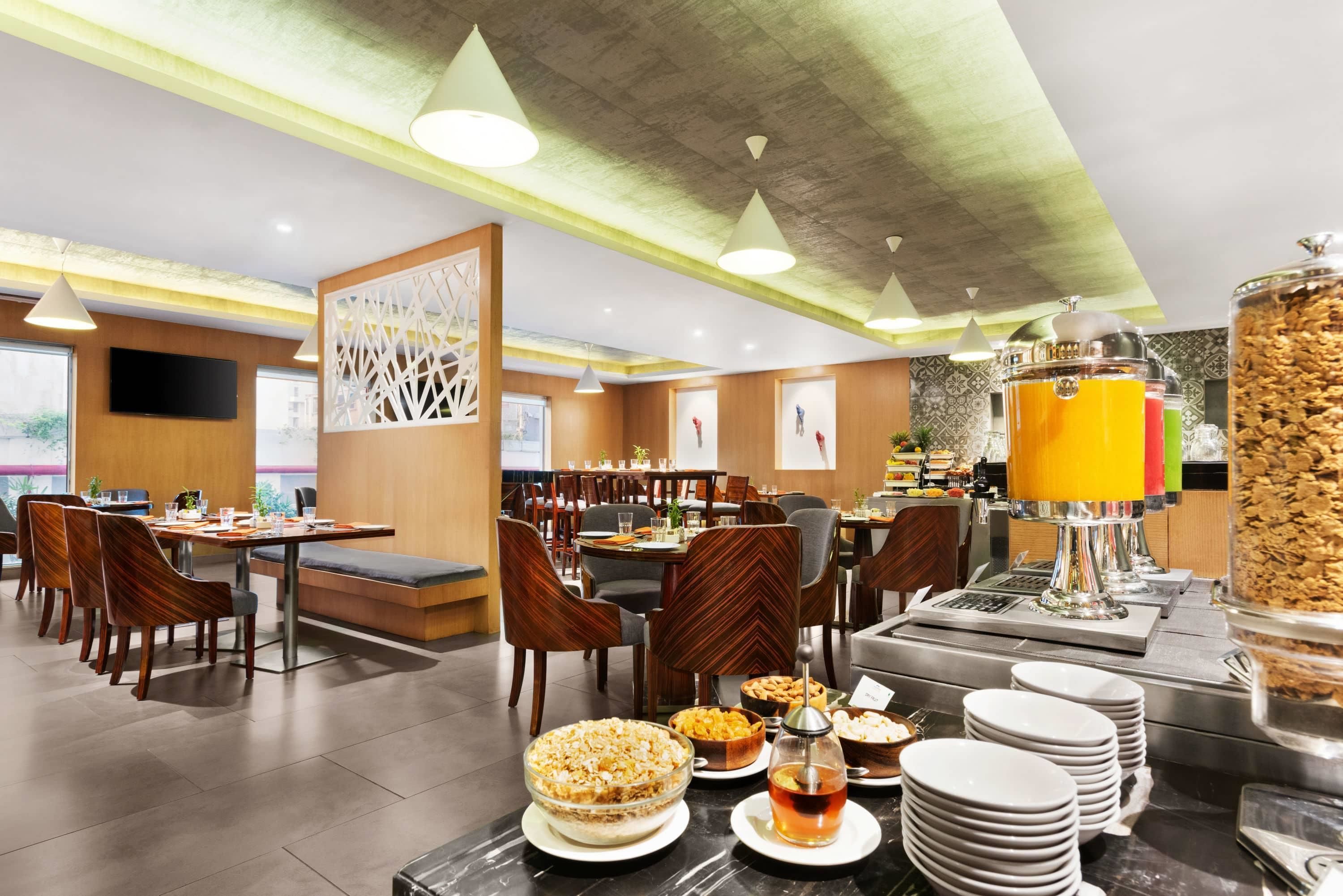 A modern dining area with multiple tables and chairs, a buffet with various food items, and a juice dispenser.
