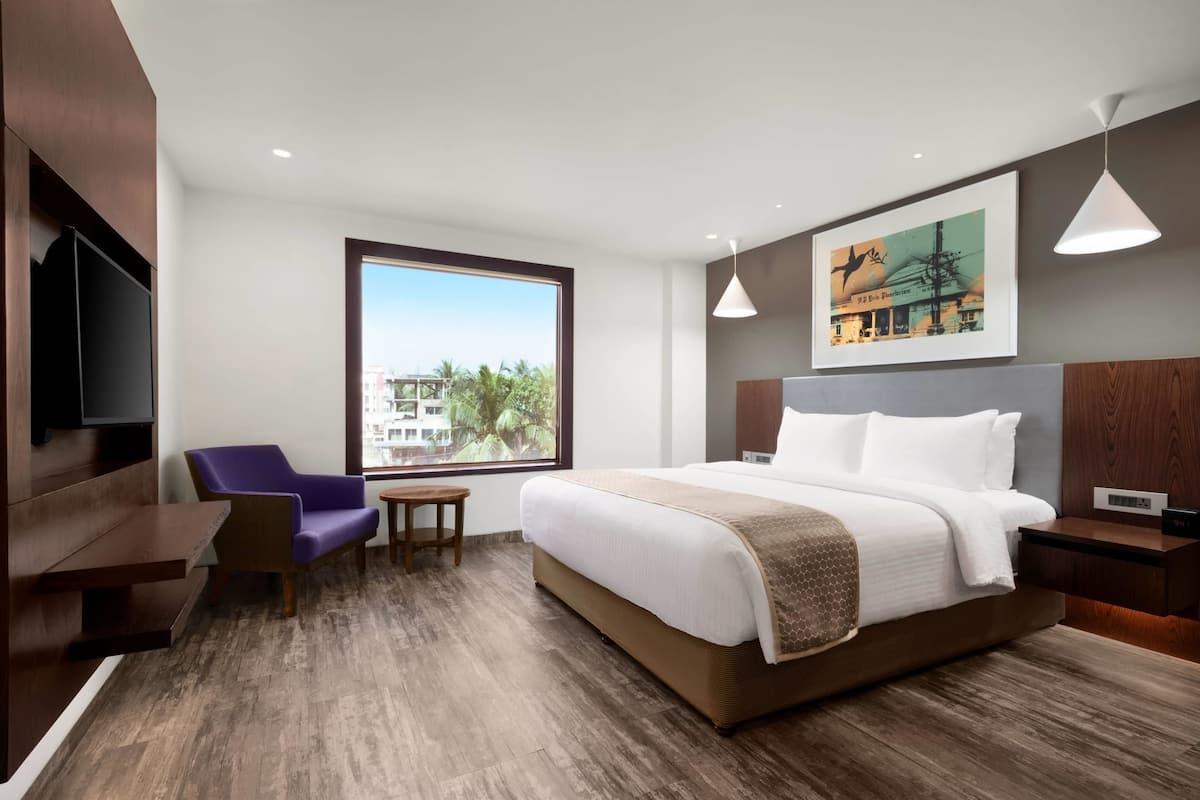Een moderne hotelkamer met een groot bed, een zithoek met een paarse stoel, een klein tafeltje en een groot raam met uitzicht op gebouwen en bomen.