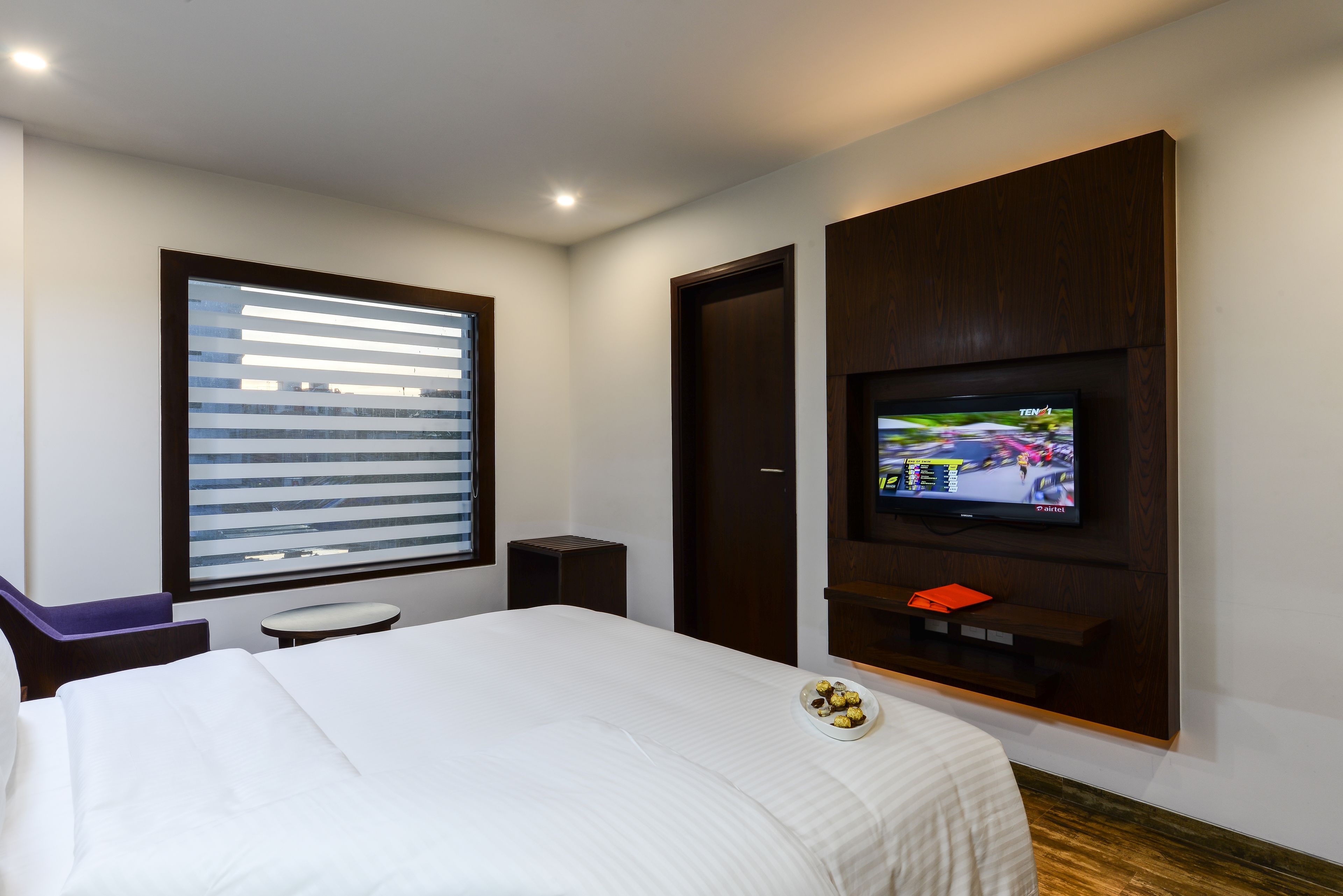 Une chambre d’hôtel avec un lit, un téléviseur fixé au mur, une fenêtre avec des stores, une petite table et une chaise.