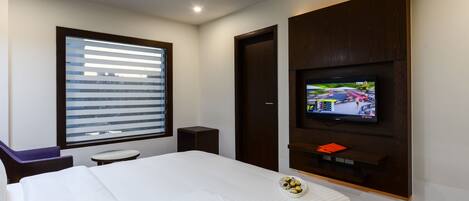 A hotel room with a bed, a TV mounted on the wall, a window with blinds, a small table, and a chair.