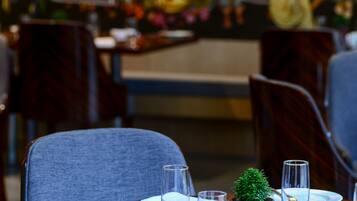 A restaurant setting with tables set for dining, a decorative mural in the background, and a small potted plant on one table.