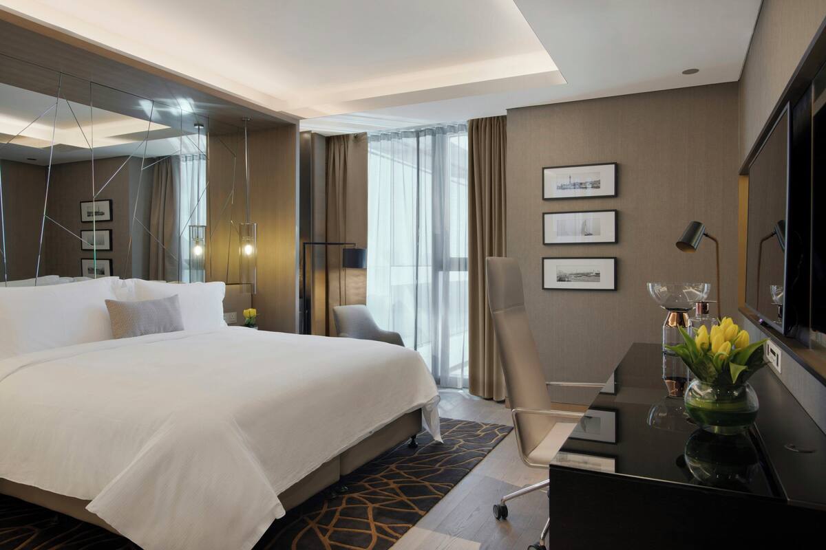 Una habitación de hotel con una cama grande, un escritorio con un florero, una silla, una lámpara de escritorio y un televisor en la pared.