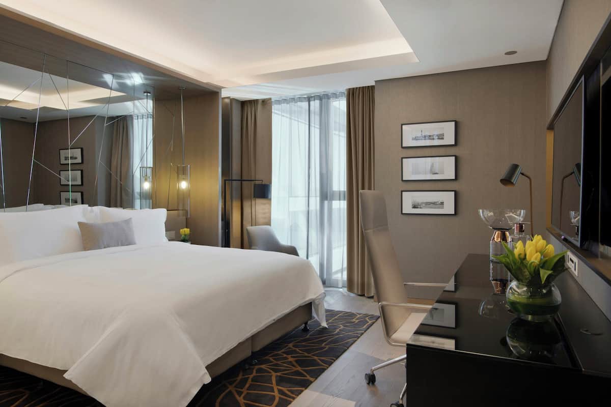 A hotel room with a large bed, a desk with a vase of flowers, a chair, a desk lamp, and a television on the wall.