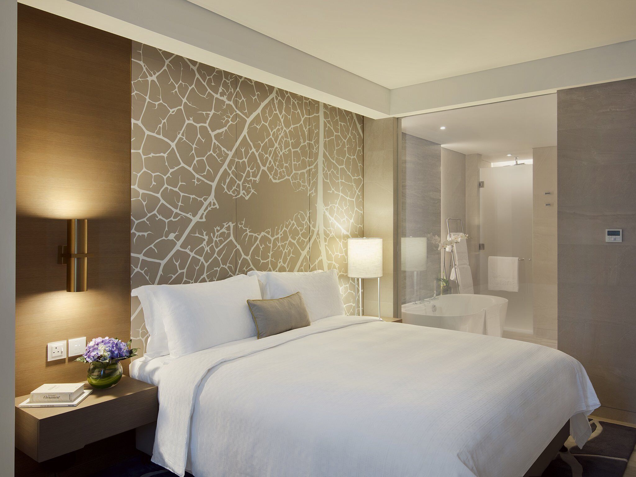 A modern hotel room with a large bed, a nightstand with a lamp, and a bathroom visible through a glass partition.