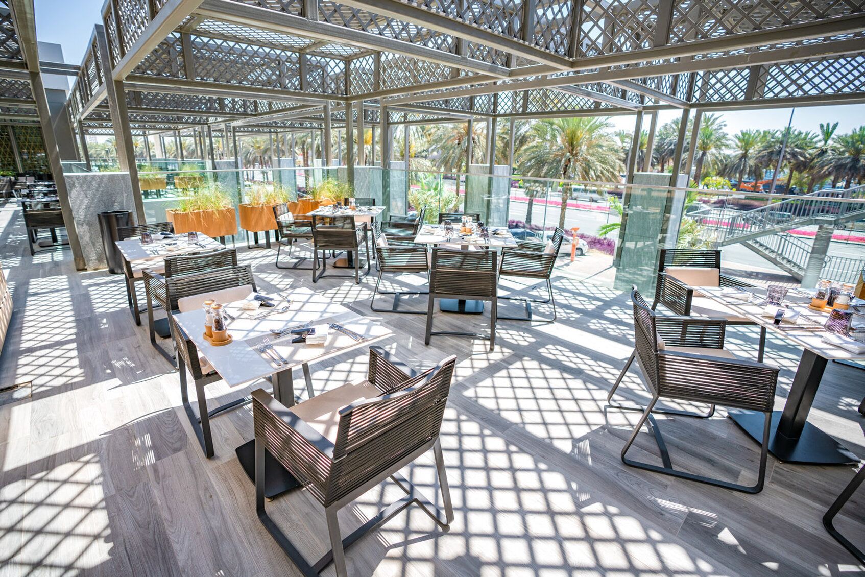 An outdoor dining area with tables and chairs, a wooden floor, and a view of palm trees and a road.