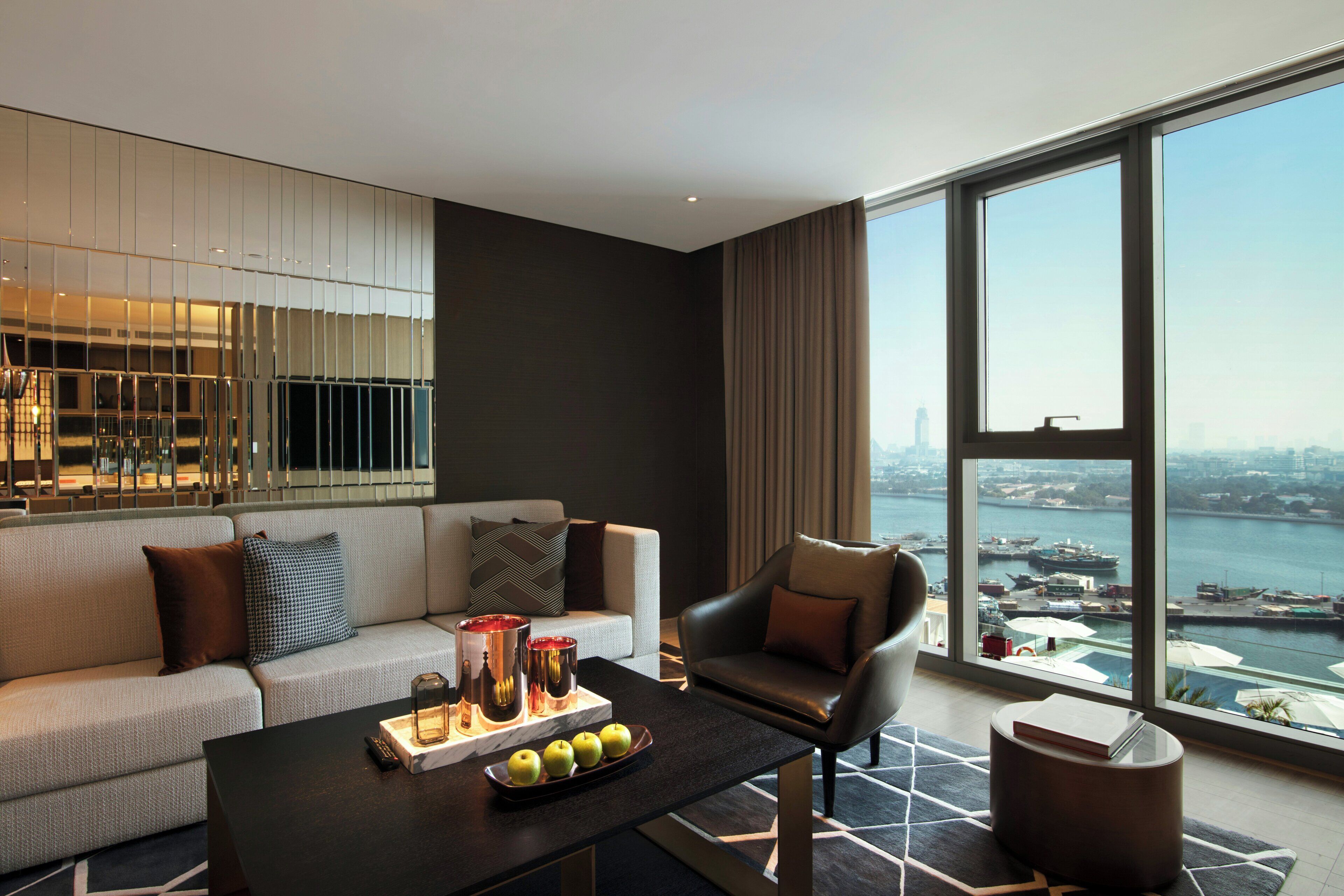 A modern hotel room with a large window, a sofa, a coffee table with fruits and candles, and a view of the cityscape.