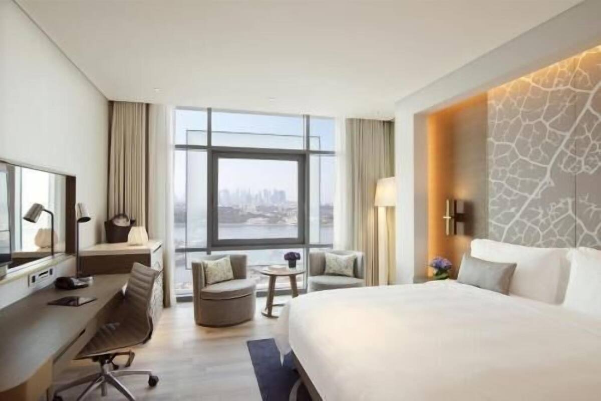 Habitación de hotel con una cama grande, un escritorio, una silla, una mesita y vistas a un paisaje urbano a través de amplias ventanas.