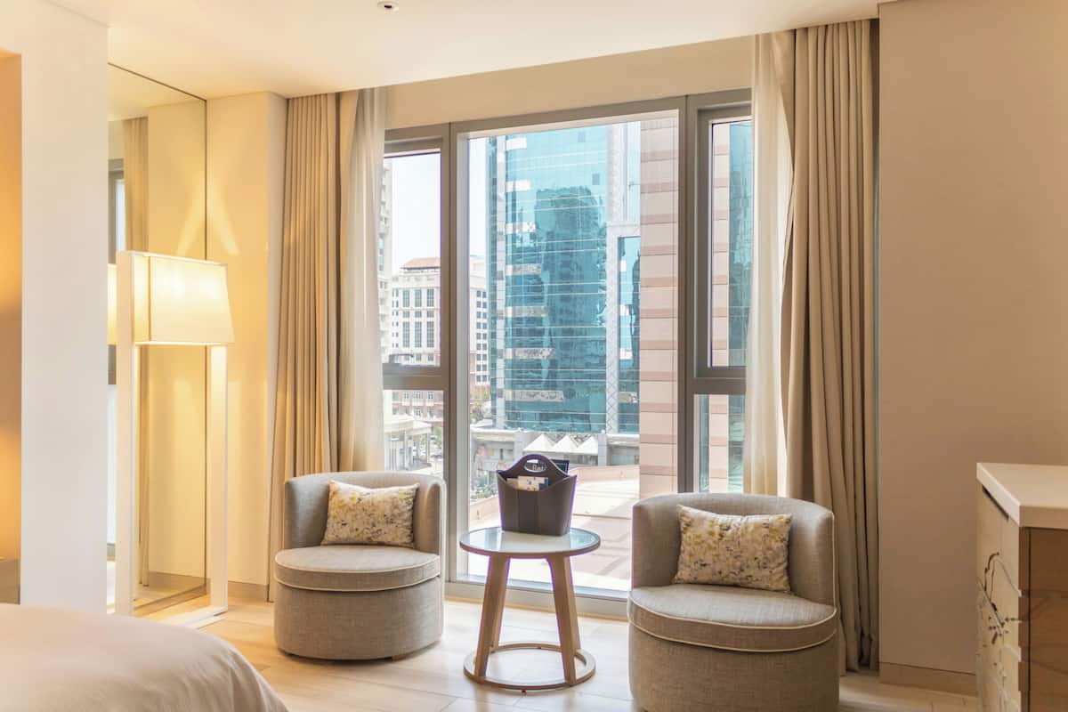 Habitación de hotel con dos sillones, una mesita, una lámpara y vistas a la ciudad a través de amplios ventanales.