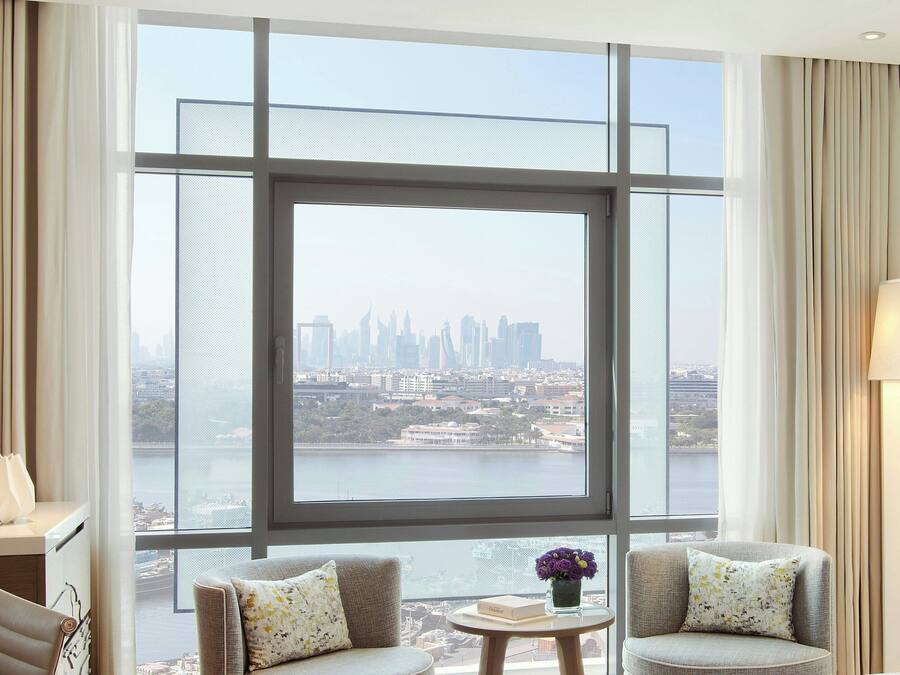 Ein modernes Hotelzimmer mit großem Fenster und Blick auf die Stadtlandschaft, einer Couch, einem kleinen Tisch und einer Lampe.