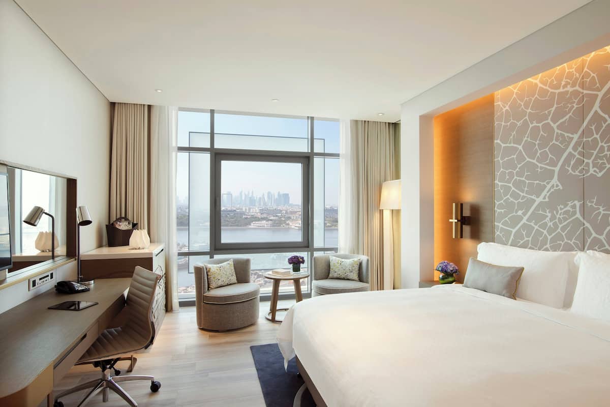 Habitación de hotel con una cama grande, un escritorio con silla, una mesita de noche con lámpara y vistas a un paisaje urbano a través de amplias ventanas.