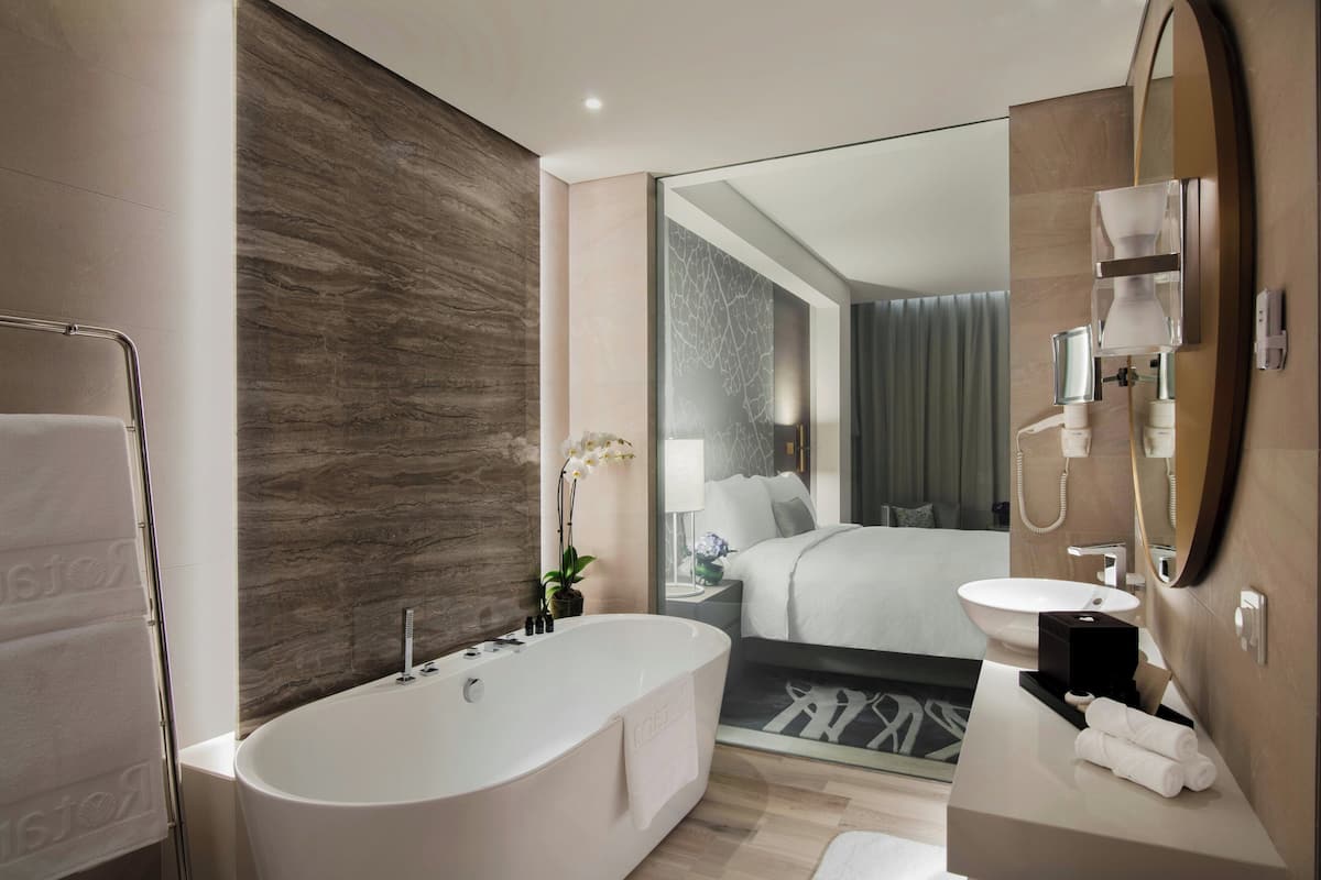 Un baño moderno con una bañera independiente, un lavabo con espejo, y un espejo grande que refleja un dormitorio con cama y cortinas.