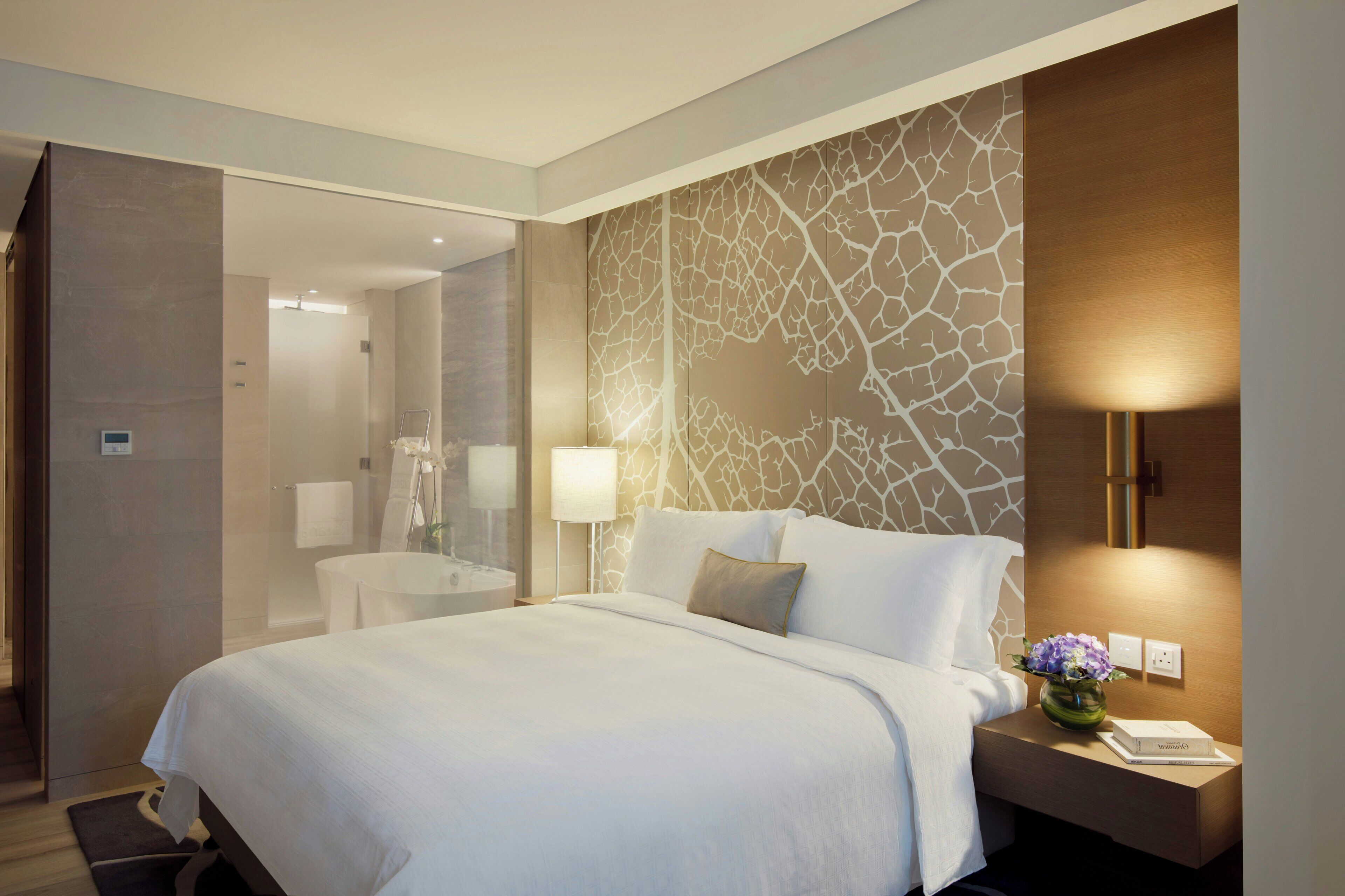 Una habitación de hotel con una cama grande, una mesita de noche con lámpara, un baño con ducha y una pared decorativa.