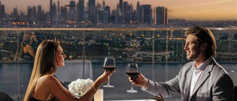 A couple toasting with wine glasses at a table with a city skyline view in the background.