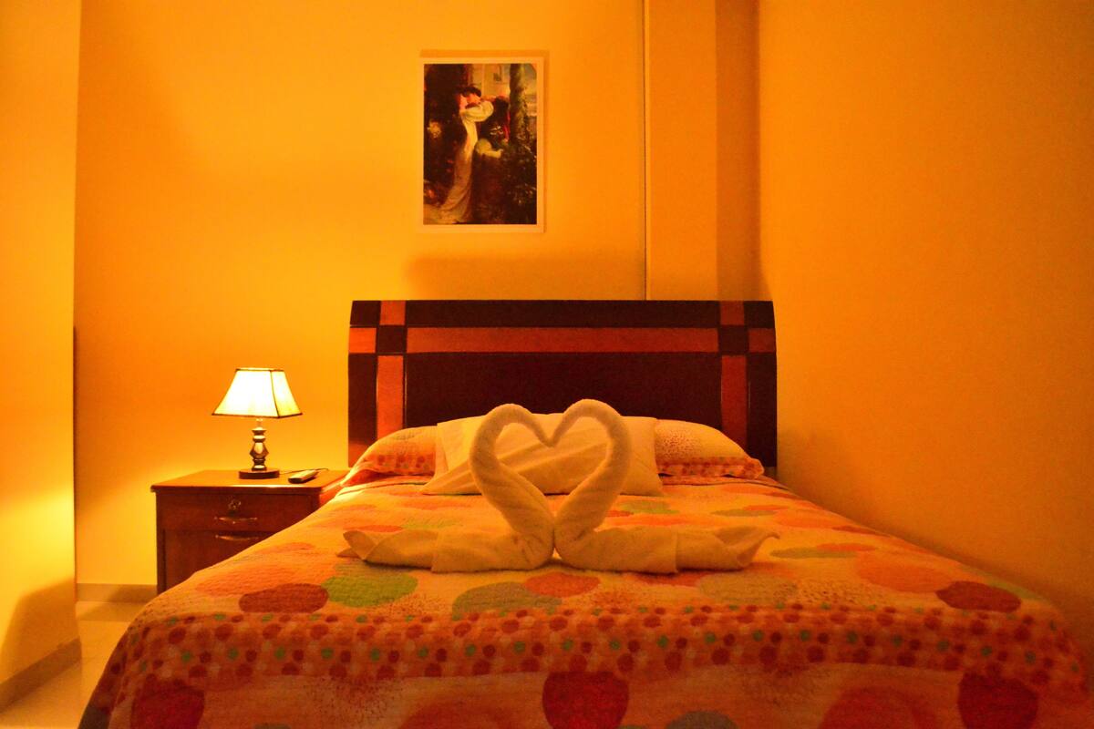 Ein Hotelzimmer mit einem Bett, das mit einer herzförmigen Handtuchgestaltung, einem Nachttisch mit Lampe und einem Bild an der Wand ausgestattet ist.