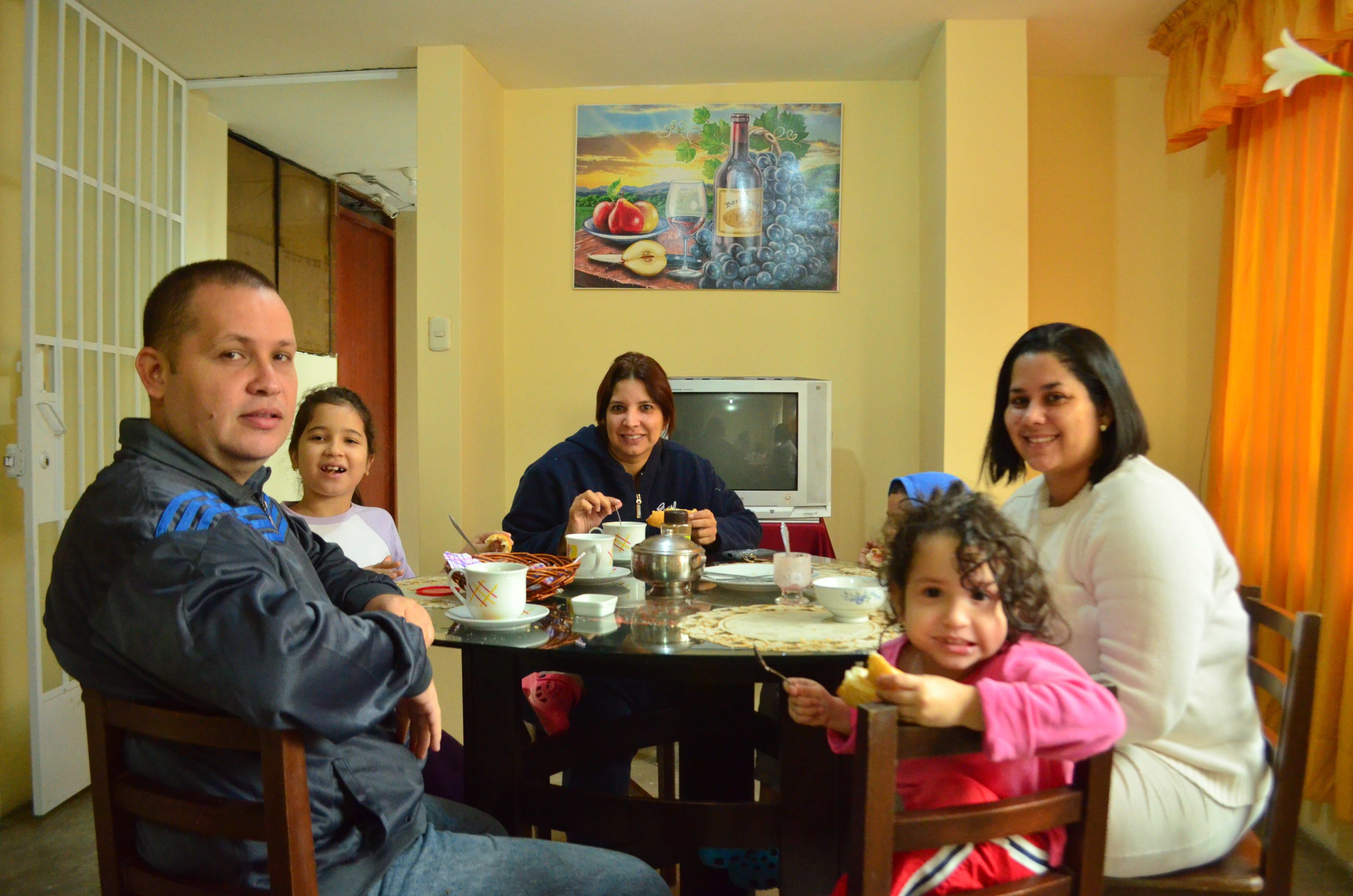 Uma família jantando à mesa, com uma pintura de uvas e vinho na parede.