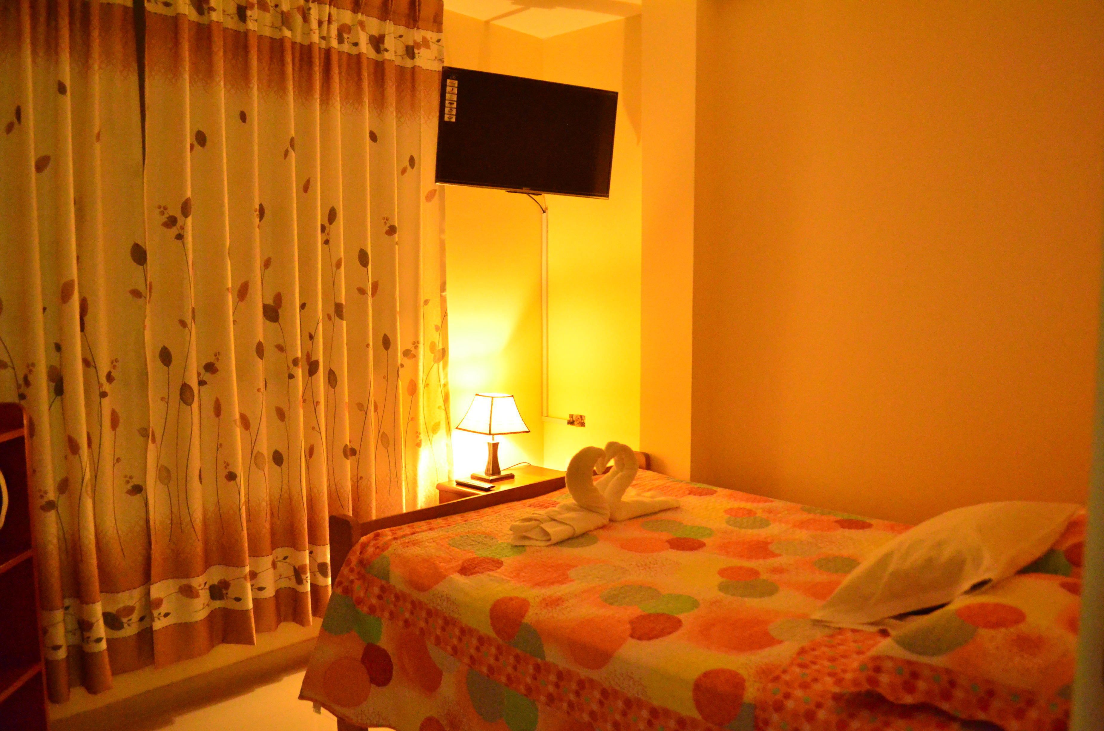 Habitación de hotel con cama, televisor de pantalla plana en la pared, lámpara en la mesita de noche y ventana con cortinas.