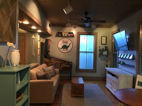 A cozy living room with a couch, a coffee table, and a television. There is a window with a view of the outside, a ceiling fan, and a wall with a dinosaur logo.
