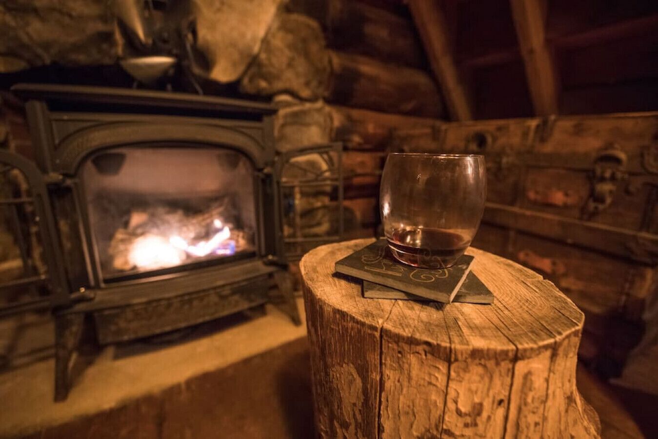 A glass of wine on a wooden coaster next to a stone fireplace with a visible fire.