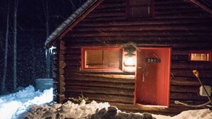 Eine Holzhütte mit roter Tür, auf der 'SKI' steht, und einem erleuchteten Fenster, umgeben von Schnee.