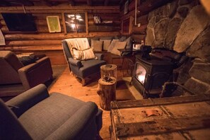 Interior acogedor de una cabaña de troncos con chimenea de piedra, un sillón azul y una mesa de madera.