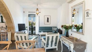Un salón acogedor con un sofá, sillas, una mesa de centro y un televisor. Hay una zona de comedor con una mesa y sillas. La habitación tiene un espejo grande y un jarrón decorativo con flores.