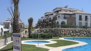 Une zone avec une piscine, un panneau indiquant les règles, un palmier et, au fond, un bâtiment doté de nombreux balcons.