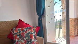 Un salon comprenant un canapé, un coussin à motifs floraux, une plante en pot et une porte-fenêtre coulissante donnant sur un espace extérieur.