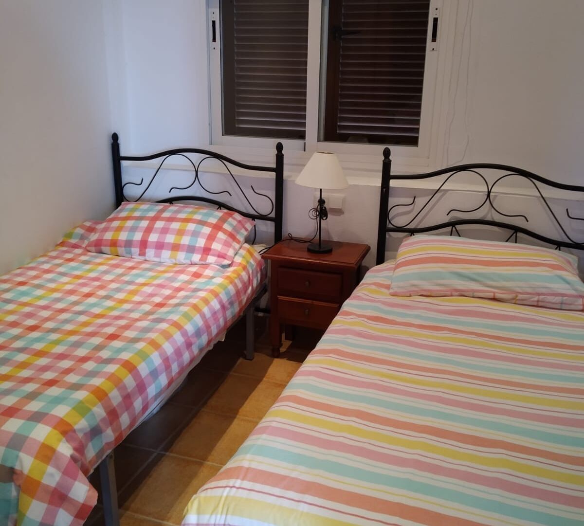 A bedroom with two single beds, a nightstand with a lamp, and a window with blinds.