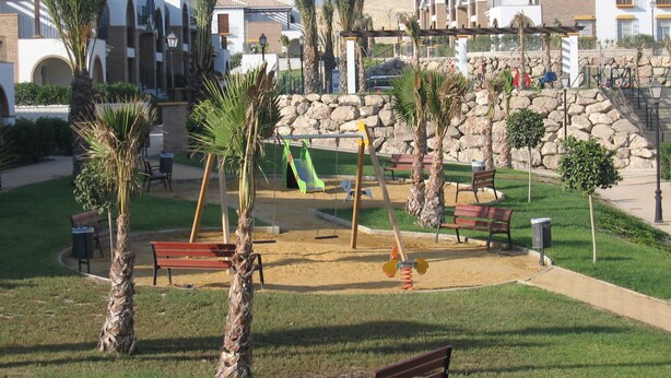Un terrain de jeux avec des balançoires et un toboggan, entouré de palmiers et de bancs, avec un mur de pierre et des bâtiments en arrière-plan.