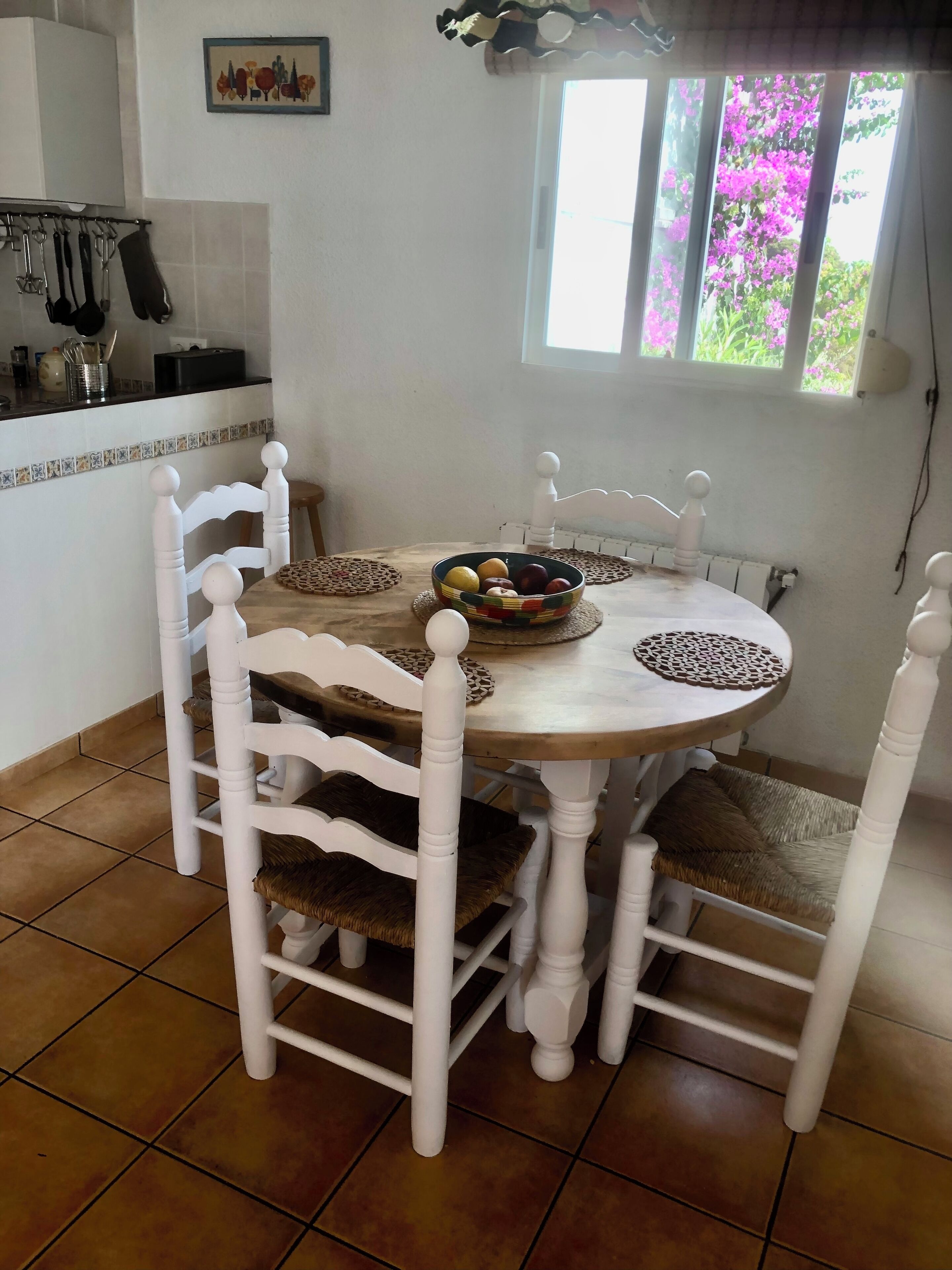 A dining area with a round wooden table, white chairs, and a bowl of fruit.