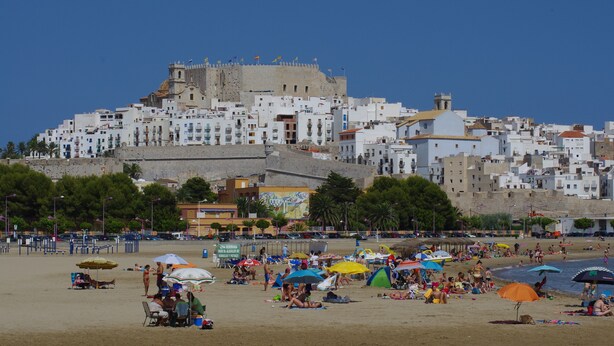 Uma praia com pessoas tomando sol e guarda-sóis, um edifício semelhante a um castelo ao fundo, e um céu azul límpido.
