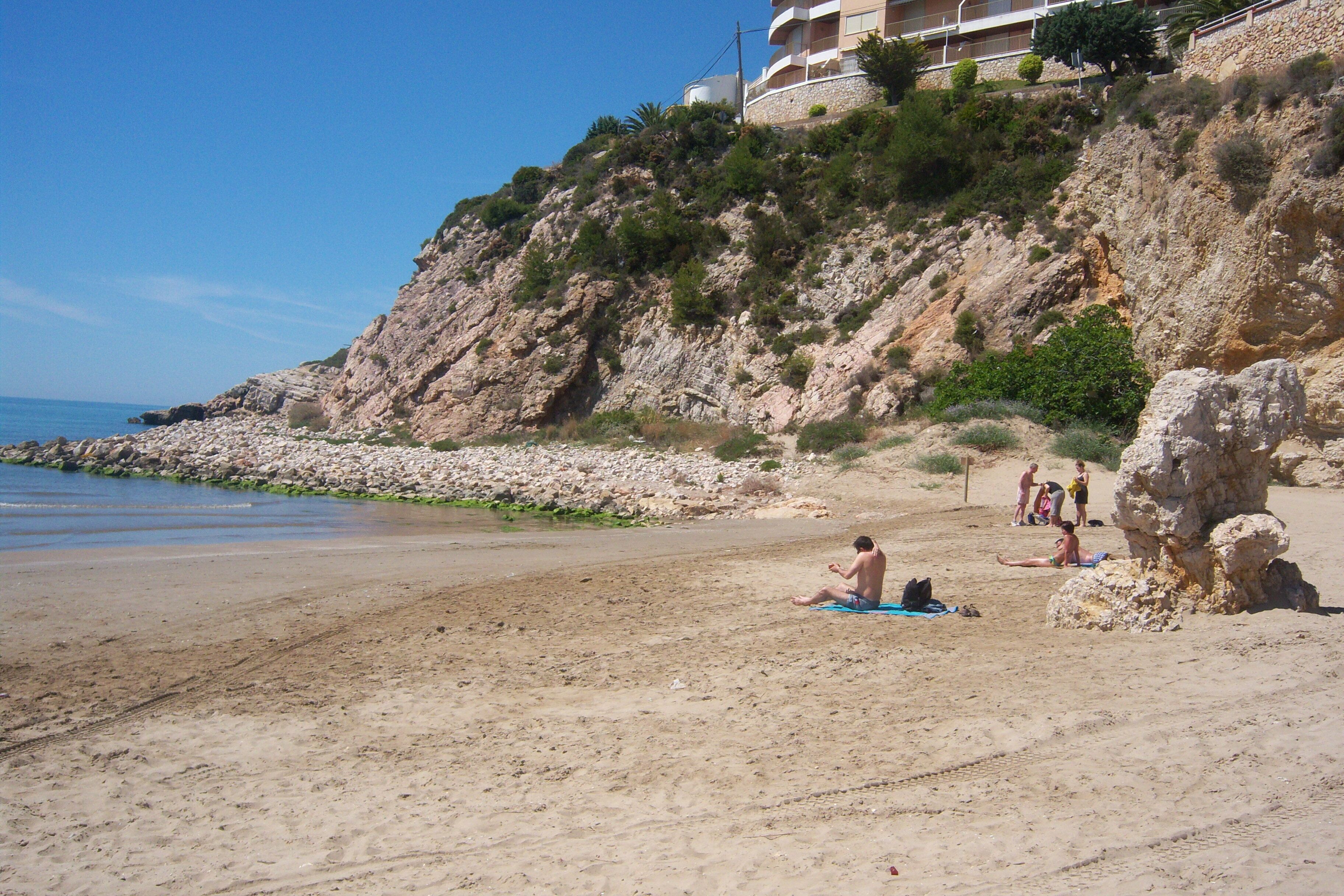 Une plage avec une falaise rocheuse, quelques personnes et un bâtiment en arrière-plan.