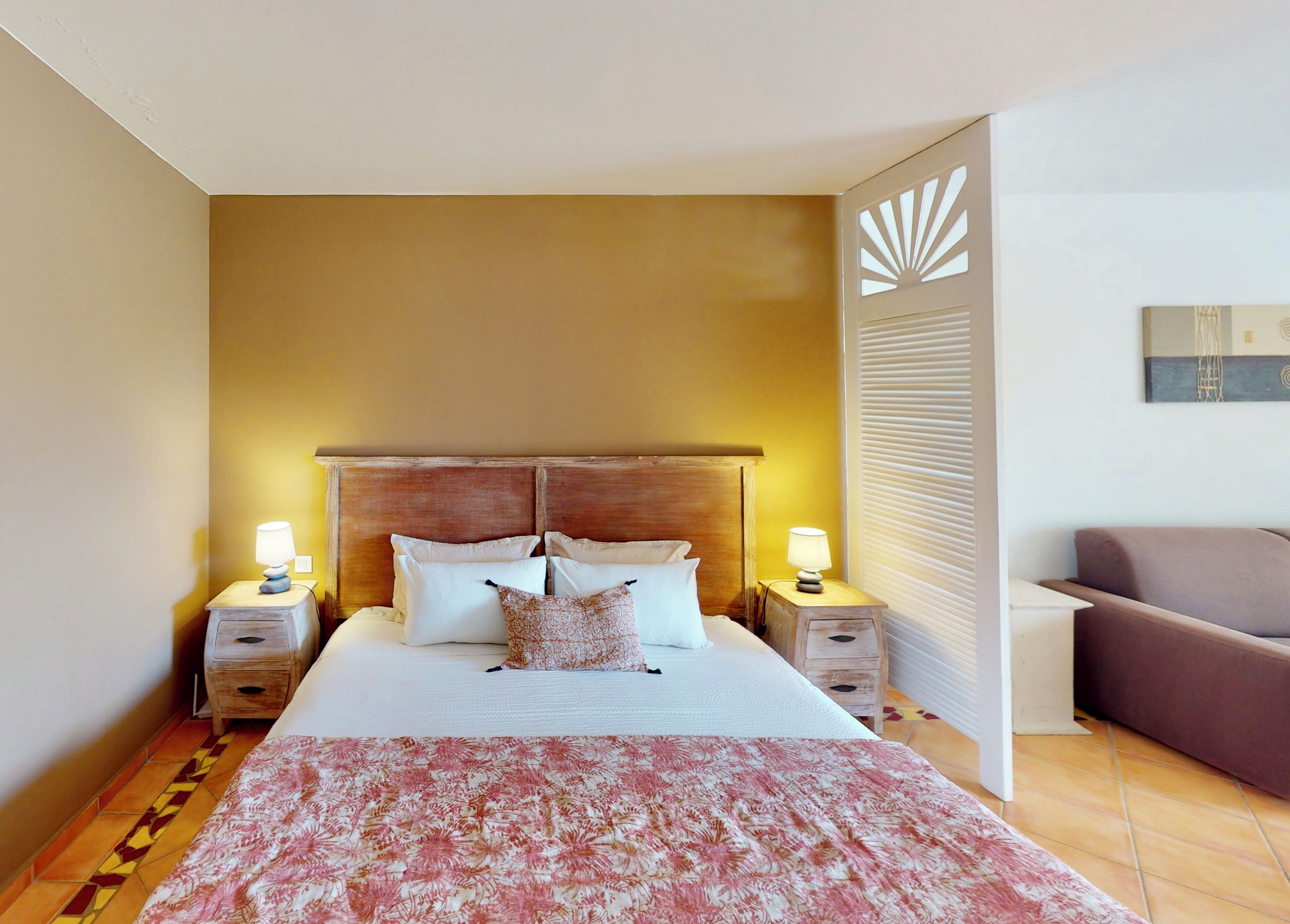 A hotel room with a bed, two bedside tables with lamps, a brown sofa, and a patterned bedspread.